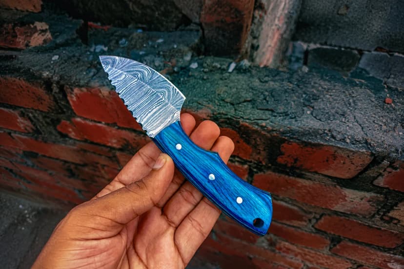 A hand holding a Damascus drop-point knife with a blue handle and a jimped spine, against a brick wall