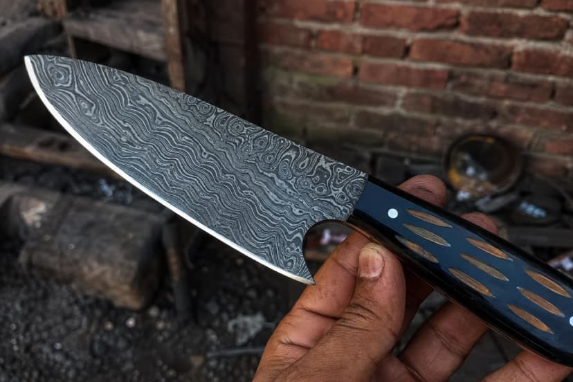A hand holding a broad Damascus blade close to the camera, the forged pattern filling the frame above a dark handle inlaid with pale ovals, a brick workshop wall behind