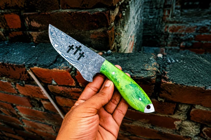 A hand holding a broad-bellied Damascus blade with three cut-outs through it and a bright green handle, against a brick wall