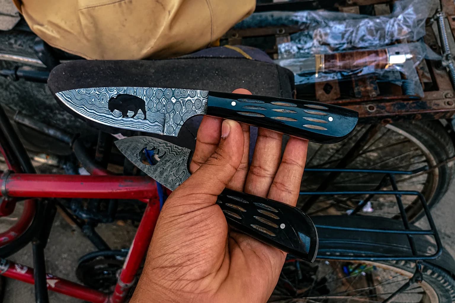 A hand holding two Damascus fixed-blade knives with dark handles inlaid with pale ovals, one blade cut through with a bison silhouette, the workshop behind them