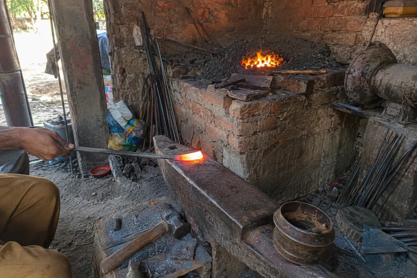 A hand holding tongs with a glowing orange billet on an anvil in a small brick forge, a coal fire burning behind it and tools scattered across the floor