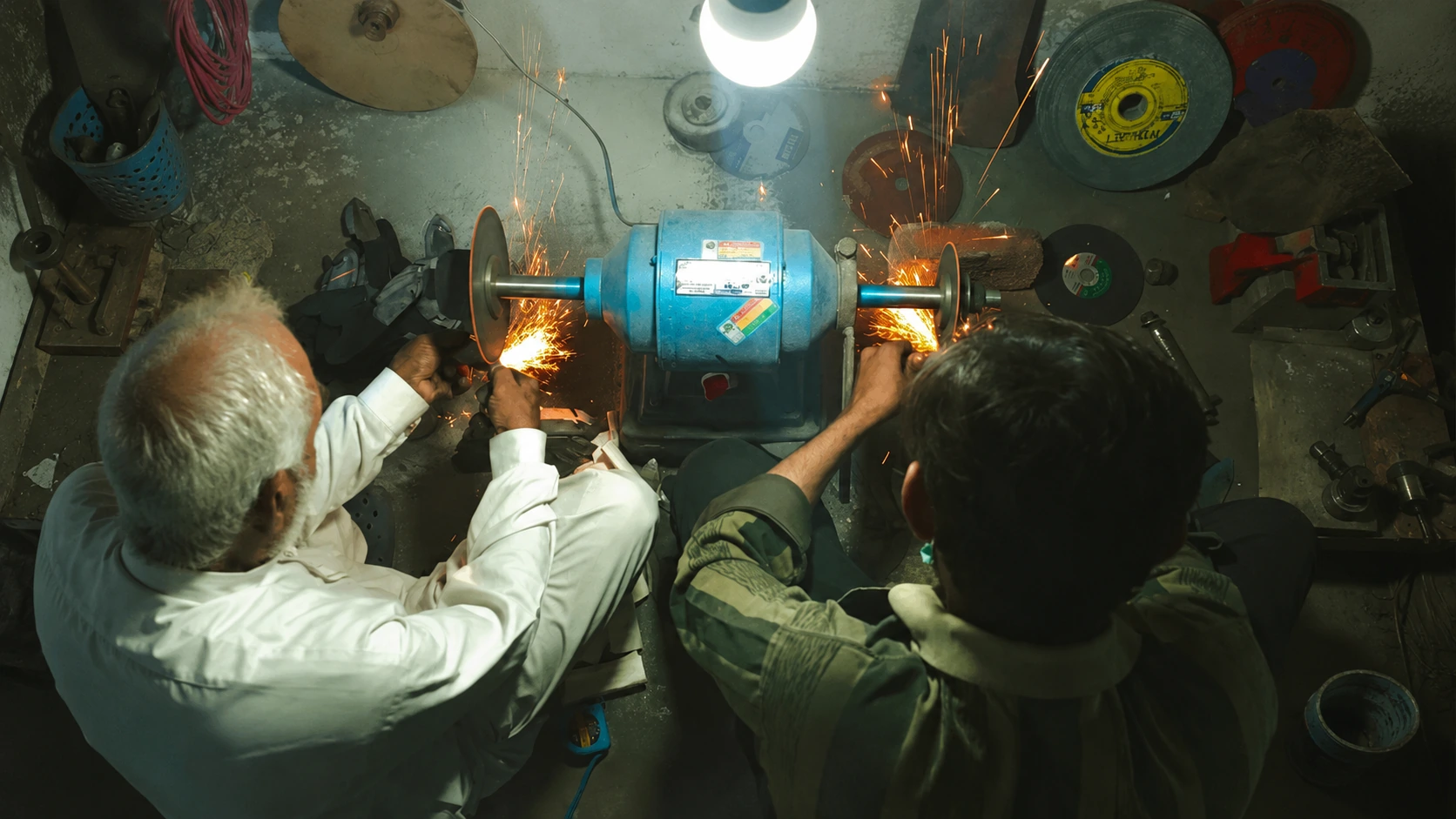 Two smiths, one grey-haired and one young, grinding blades side by side at a twin bench grinder